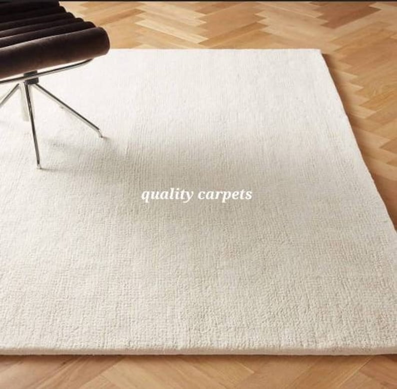 May include: A rectangular off-white area rug with a textured, woven surface. The rug features the text quality carpets printed in white in the center. A portion of a dark brown chair with a chrome metal base is visible in the upper left corner.