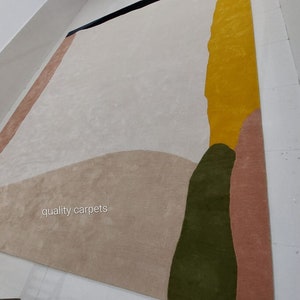 May include: Abstract area rug with a beige background, featuring large shapes in shades of pink, green, and yellow. The text "quality carpets" is visible in the lower left corner.