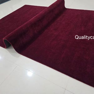 May include: A rectangular area rug in a deep burgundy color with a soft, plush texture. The rug is partially folded over, revealing a dark underside. The text "Qualitycarpets" is printed in white on the right side of the rug surface.