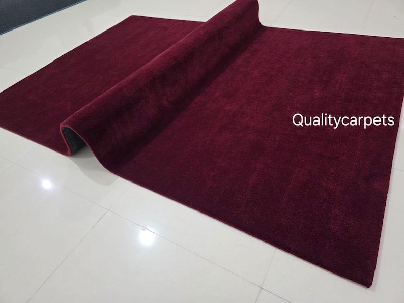 May include: A rectangular area rug in a deep burgundy color with a soft, plush texture. The rug is partially folded over, revealing a dark underside. The text "Qualitycarpets" is printed in white on the right side of the rug surface.