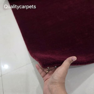 May include: A deep burgundy, solid-colored rug with a soft, plush texture. The corner of the rug is lifted to reveal its thickness and pile. The text "Qualitycarpets" is printed in white in the upper left corner of the image.