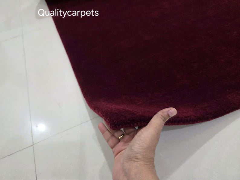 May include: A deep burgundy, solid-colored rug with a soft, plush texture. The corner of the rug is lifted to reveal its thickness and pile. The text "Qualitycarpets" is printed in white in the upper left corner of the image.
