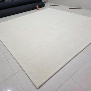 May include: A large, square, solid off-white area rug with a smooth, uniform texture. The rug features clean, straight edges and a flat pile surface, presenting a neutral and consistent color throughout its entire rectangular expanse.