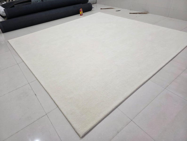 May include: A large, square, solid off-white area rug with a smooth, uniform texture. The rug features clean, straight edges and a flat pile surface, presenting a neutral and consistent color throughout its entire rectangular expanse.