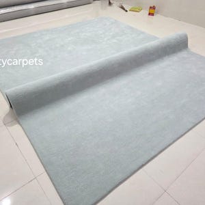 May include: A large, rectangular, light blue area rug with a soft, uniform texture. The rug is partially rolled over itself, revealing a consistent solid color throughout. The text Qualitycarpets is printed in white on the left side of the rug.
