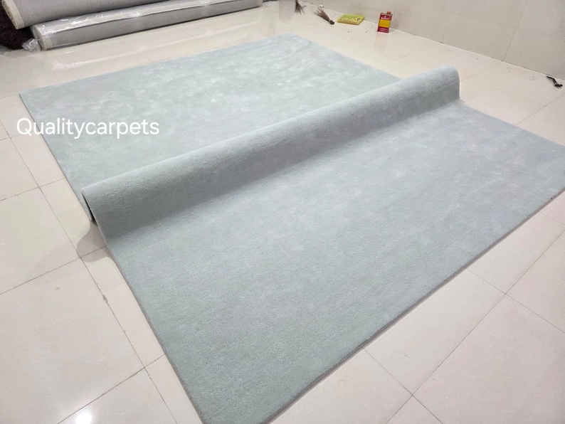 May include: A large, rectangular, light blue area rug with a soft, uniform texture. The rug is partially rolled over itself, revealing a consistent solid color throughout. The text Qualitycarpets is printed in white on the left side of the rug.