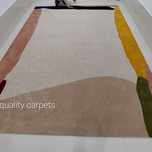 May include: A large, abstract area rug with a cream background and colourful geometric shapes in shades of pink, yellow, green, and blue. The rug has the text "quality carpets" printed on it.