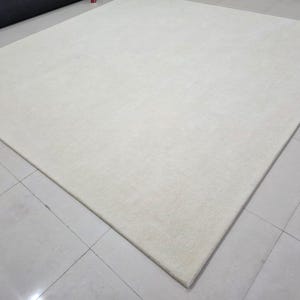 May include: A large, square-shaped area rug in a solid, off-white color. The rug features a dense, short-pile texture with a clean, finished edge. The surface appears uniform and smooth, covering a significant portion of the floor space shown.