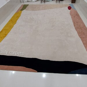 May include: Abstract area rug with a cream background and geometric shapes in yellow, green, blue, red, black, and pink. The rug has the text "quality carpets" in the bottom left corner.