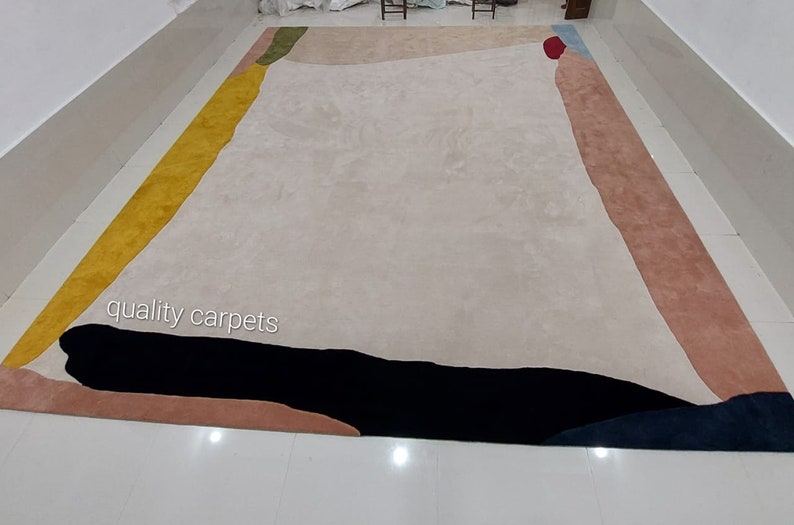 May include: Abstract area rug with a cream background and geometric shapes in yellow, green, blue, red, black, and pink. The rug has the text "quality carpets" in the bottom left corner.