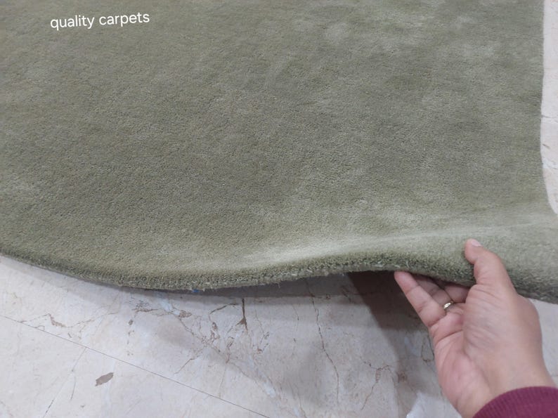 May include: A sage green carpet with a thick, plush pile texture. The surface is uniform in color and features a soft, dense appearance. The text "quality carpets" is printed in white in the upper left corner of the image.