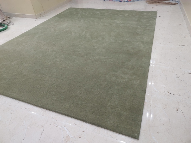 May include: A rectangular area rug in a solid sage green color with a short, dense pile texture. The rug features clean, straight edges and a uniform appearance across its entire surface, lying flat on a light-colored, reflective floor.