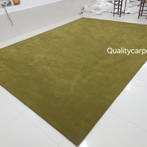 May include: A large, rectangular area rug in a solid olive green color with a soft, uniform texture. The text "Qualitycarpets" is printed in white on the surface of the rug. The rug is displayed flat on a white tiled floor.