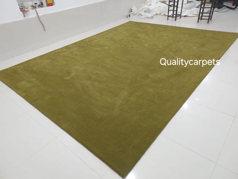 May include: A large, rectangular area rug in a solid olive green color with a soft, uniform texture. The text "Qualitycarpets" is printed in white on the surface of the rug. The rug is displayed flat on a white tiled floor.