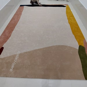 May include: Abstract area rug with a beige background and colourful geometric shapes in shades of pink, yellow, green, blue, and brown. The text "quality carpets" is printed on the rug.