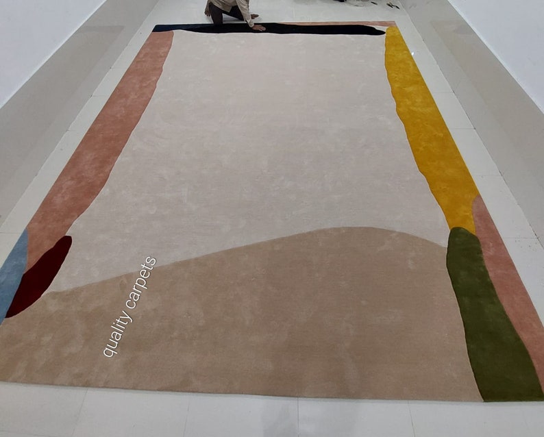 May include: Abstract area rug with a beige background and colourful geometric shapes in shades of pink, yellow, green, blue, and brown. The text "quality carpets" is printed on the rug.