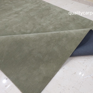 May include: A solid sage green area rug with a soft, uniform texture. One corner of the rug is folded over to reveal a dark gray backing. The text qualitycarpets is visible in the upper right corner of the image against the rug surface.