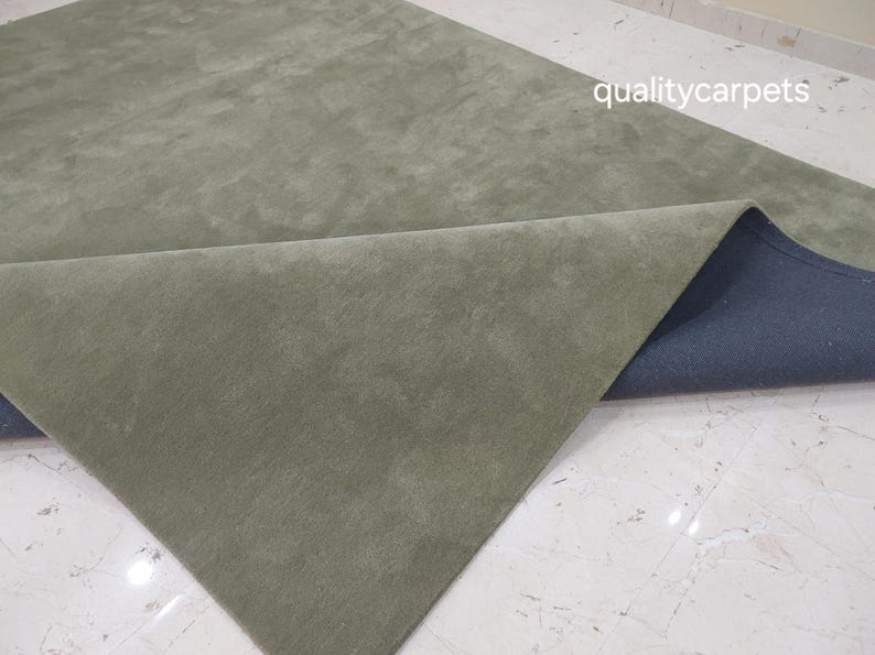 May include: A solid sage green area rug with a soft, uniform texture. One corner of the rug is folded over to reveal a dark gray backing. The text qualitycarpets is visible in the upper right corner of the image against the rug surface.