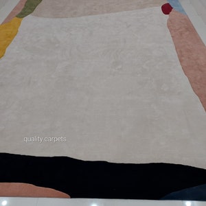 May include: A modern abstract area rug with a white background and geometric shapes in shades of pink, yellow, green, blue, and black. The rug has the text "quality carpets" in the centre.