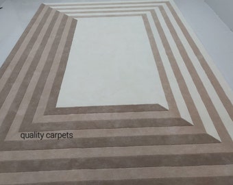 May include: A beige and white area rug with a geometric pattern. The rug has a large, white rectangle in the centre surrounded by a series of beige stripes. The text "quality carpets" is visible in the bottom left corner of the rug.