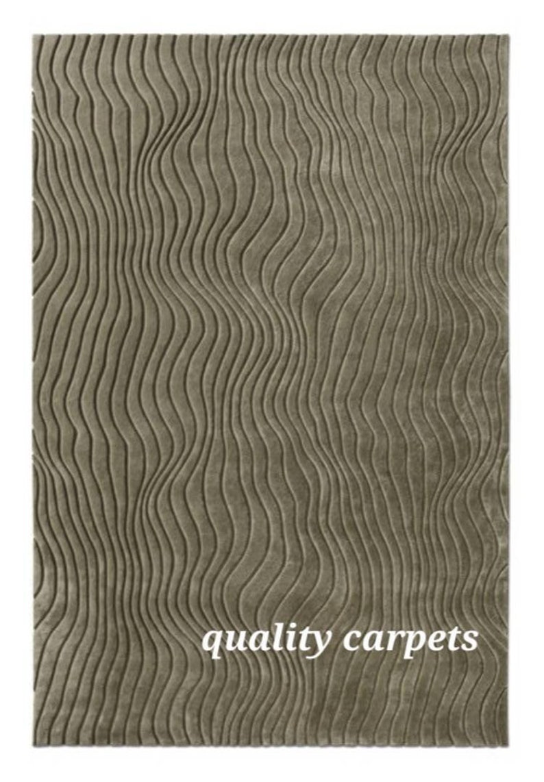 May include: A close-up of a light brown rug with a wavy, textured pattern. The rug has a subtle sheen and a soft, plush feel. The text "quality carpets" is printed in the lower right corner of the rug.