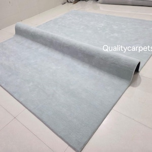May include: A large, rectangular, light gray area rug with a soft, uniform texture. The rug is partially folded over itself, revealing its thickness and backing. The text "Qualitycarpets" is printed in white on the surface of the rug.