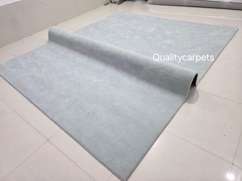 May include: A large, rectangular, light gray area rug with a soft, uniform texture. The rug is partially folded over itself, revealing its thickness and backing. The text "Qualitycarpets" is printed in white on the surface of the rug.