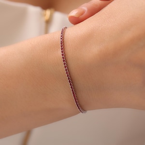 Puede incluir: Una delicada pulsera de plata adornada con una línea continua de pequeñas y vibrantes piedras preciosas rojas. La pulsera se muestra en la muñeca de una persona, destacando su diseño elegante y refinado. Esta joya es adecuada para el uso diario o para ocasiones especiales.