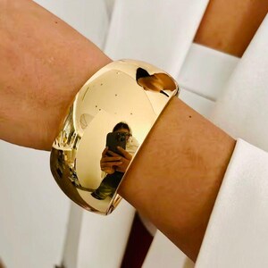 Puede incluir: Un brazalete ancho y pulido en tono dorado. El brazalete tiene una superficie lisa y reflectante, con un dise&ntilde;o curvo. La superficie refleja el entorno, incluida una persona que sostiene un tel&eacute;fono. El brazalete se lleva en la mu&ntilde;eca.