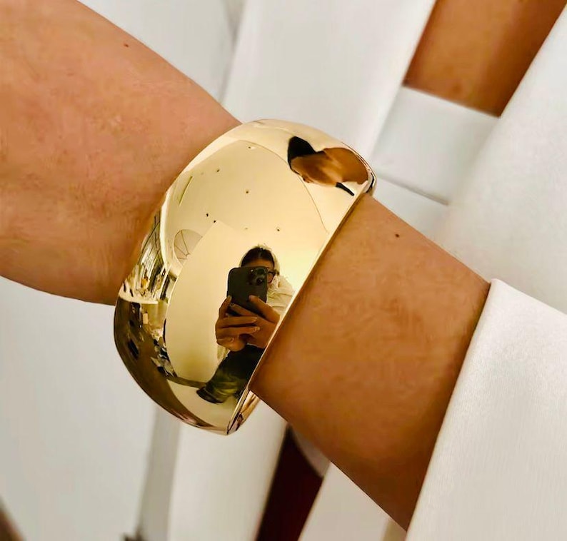 Puede incluir: Un brazalete ancho y pulido en tono dorado. El brazalete tiene una superficie lisa y reflectante, con un dise&ntilde;o curvo. La superficie refleja el entorno, incluida una persona que sostiene un tel&eacute;fono. El brazalete se lleva en la mu&ntilde;eca.
