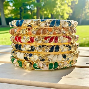 May include: A stack of six gold-toned bangles with colorful enamel designs. Each bangle features a different color scheme, including blue, orange, red, black, white, and green, accented with clear crystals. The bangles are stacked on a light-colored wooden surface.