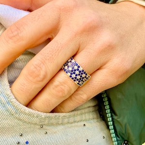 May include: A gold-toned ring with a wide band, encrusted with small, sparkling blue and white stones. The ring is shown on a finger, with a denim pocket and green jacket in the background. The stones are arranged in a repeating pattern.
