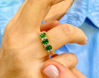 May include: A gold ring featuring a row of emerald-cut green gemstones and small clear stones. The ring has a textured, woven-like band. The ring is shown on a person's finger against a light blue background.