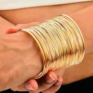 May include: A stack of gold-colored bangles. The bangles are made of thin, shiny metal and are stacked together to create a wide cuff bracelet. The bracelets are worn on the wrist.