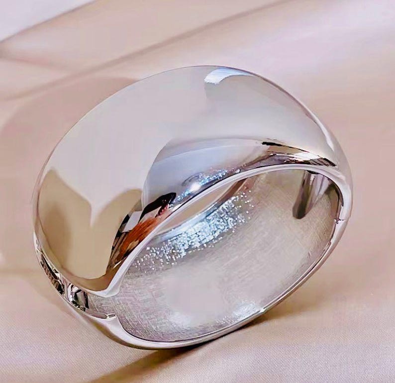 Puede incluir: Una pulsera ancha y pulida de plata con un dise&ntilde;o redondeado y abovedado. La pulsera refleja la luz, creando una apariencia brillante y reluciente. La joya es un accesorio llamativo.