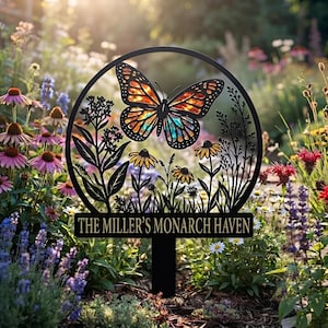 May include: A black metal garden stake featuring a circular frame with a monarch butterfly and wildflowers. The text THE MILLER'S MONARCH HAVEN is displayed on a horizontal bar at the bottom of the circular design, which is set in a garden.