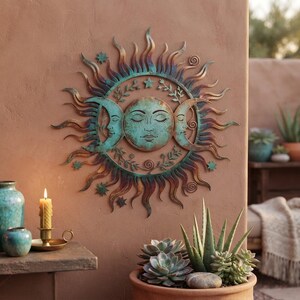 May include: A circular metal wall art piece featuring a central sun face flanked by two crescent moon faces. The design includes radiating sunbeams, small stars, and leafy vine accents in a weathered teal and copper patina finish.