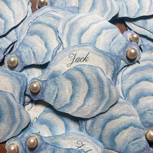 May include: A collection of watercolor oyster shell place cards. Each card is a light blue and white with a pearl accent. The name "Jack" is written on one of the cards in elegant script. These cards are ideal for a nautical themed event.