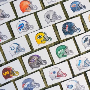 Peut inclure: Illustrations à l'aquarelle de divers casques de football américain de la NFL sur des cartes blanches. Les casques présentent les couleurs et les logos des équipes, notamment les Cowboys de Dallas, les Dolphins de Miami et les Patriots de la Nouvelle-Angleterre. Les cartes sont disposées sur une surface verte texturée.