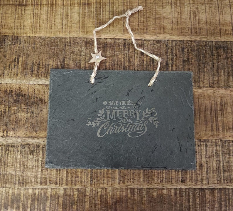May include: A black slate sign with a white inscription that reads "Have yourself a merry little Christmas". The sign is hanging from a string with a small star-shaped ornament.