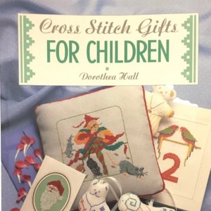 May include: A book titled "Cross Stitch Gifts For Children" by Dorothea Hall. The cover features a variety of cross stitch projects, including a cushion with a jester design, a small stuffed toy, and a card with a Santa Claus design.