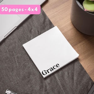 Może przedstawiać: Biały kwadratowy notatnik z czarnym napisem "Grace". Notatnik ma wymiary 10x10 cm i zawiera 50 stron. Notatnik leży na ciemnoszarej powierzchni, w tle laptop i roślina doniczkowa.