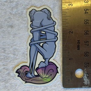 Puede incluir: Una pegatina con una ilustraci&oacute;n estilizada de dos figuras entrelazadas. Las figuras est&aacute;n representadas en tonos azules, con un dise&ntilde;o arremolinado y colorido en la base. La pegatina mide aproximadamente 7,6 cm de altura.
