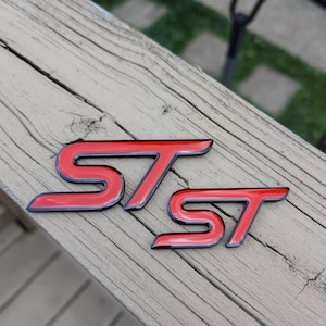 May include: Two automotive emblems featuring the letters ST in a bold, stylized font. Each emblem has a vibrant red center inlay surrounded by a thin, raised black border, creating a high-contrast appearance for these vehicle exterior accessories.