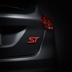 May include: A dark gray vehicle trunk featuring a red ST emblem with a carbon fiber texture. The emblem is positioned on the right side of the trunk lid, adjacent to a red and clear taillight assembly, set against a dark, shadowed background.