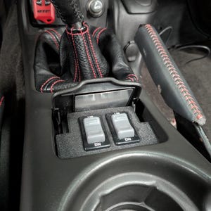 May include: Close-up of a car's center console with two gray switches labeled "DN" and "UP". The console is black with red stitching and a black leather gear shifter.