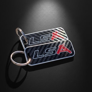 May include: Two rectangular keychains featuring a black and grey carbon fiber pattern. Each keychain displays the white outlined text LS and a red letter A. A silver metal split ring is attached to the top corner of each rectangular tag.