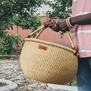 May include: A woven basket with a rounded shape and two handles. The basket is tan with brown leather accents on the handles. A small leather tag on the front reads "TAILORED COLLECTION".