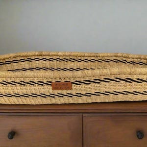 May include: A woven oval basket with black and natural colored stripes and leather handles. The basket has a label that reads "Tailored Collection".