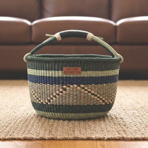 May include: A round woven basket with a handle. The basket features green and blue horizontal stripes and a woven pattern at the bottom. A small rectangular tag reads "TAYLORED COLLECTION".